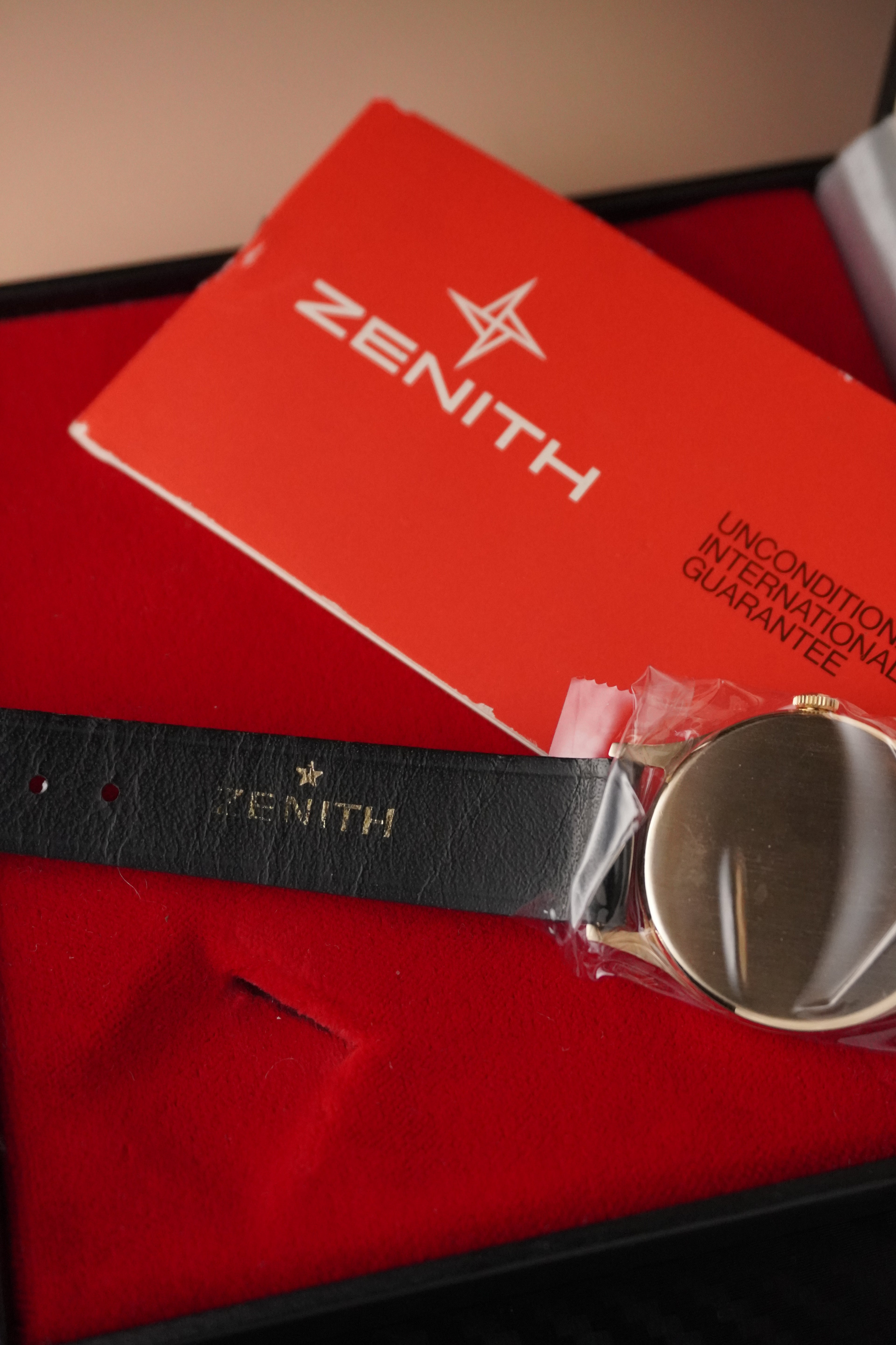 Gold watch with black strap and 'ZENITH' branding on a red cushion