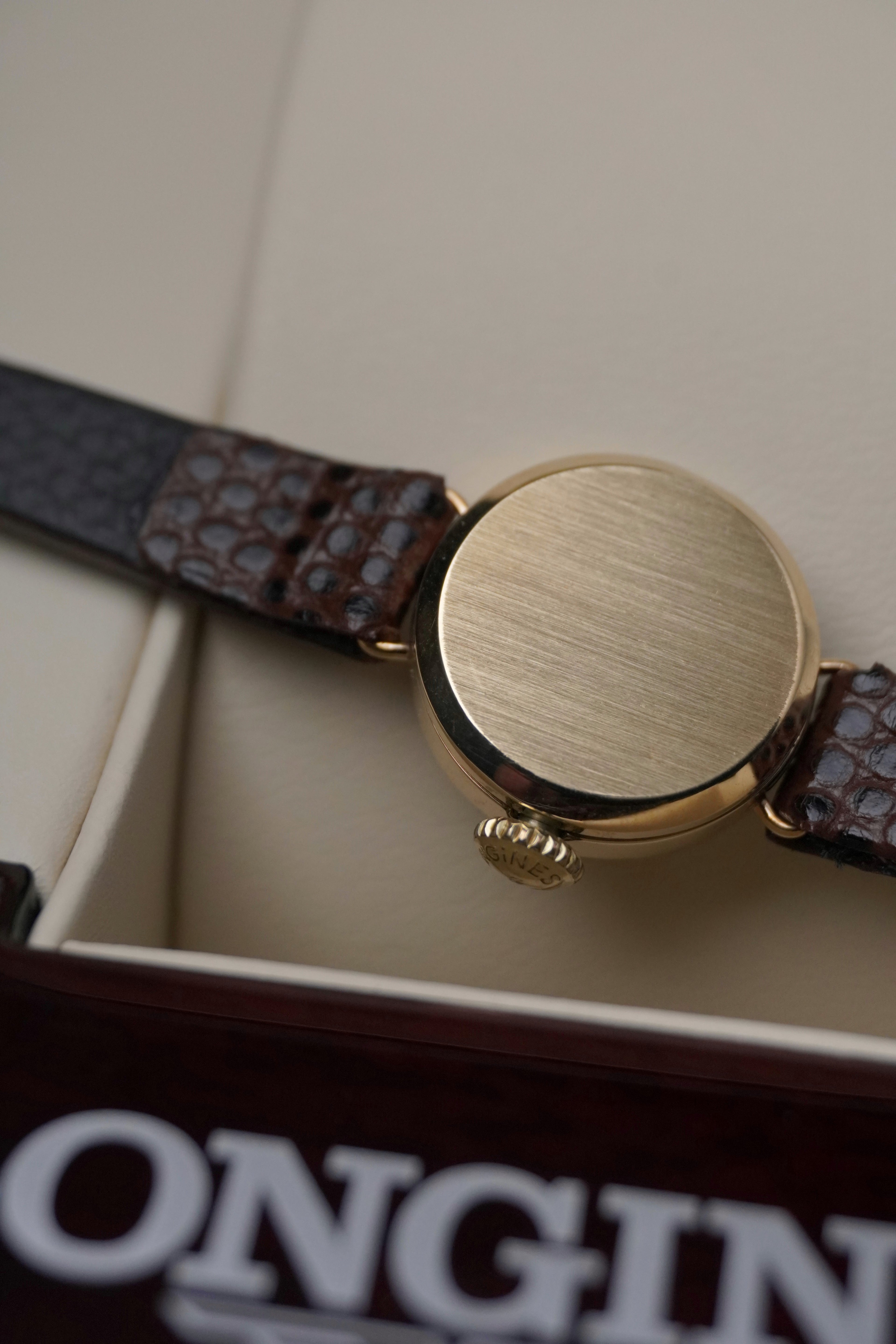 
Vintage Longines ladies’ wristwatch with a textured gold round case, white dial, slim gold hour markers, and brown leather strap, displayed on a cream watch cushion inside a Longines presentation box.