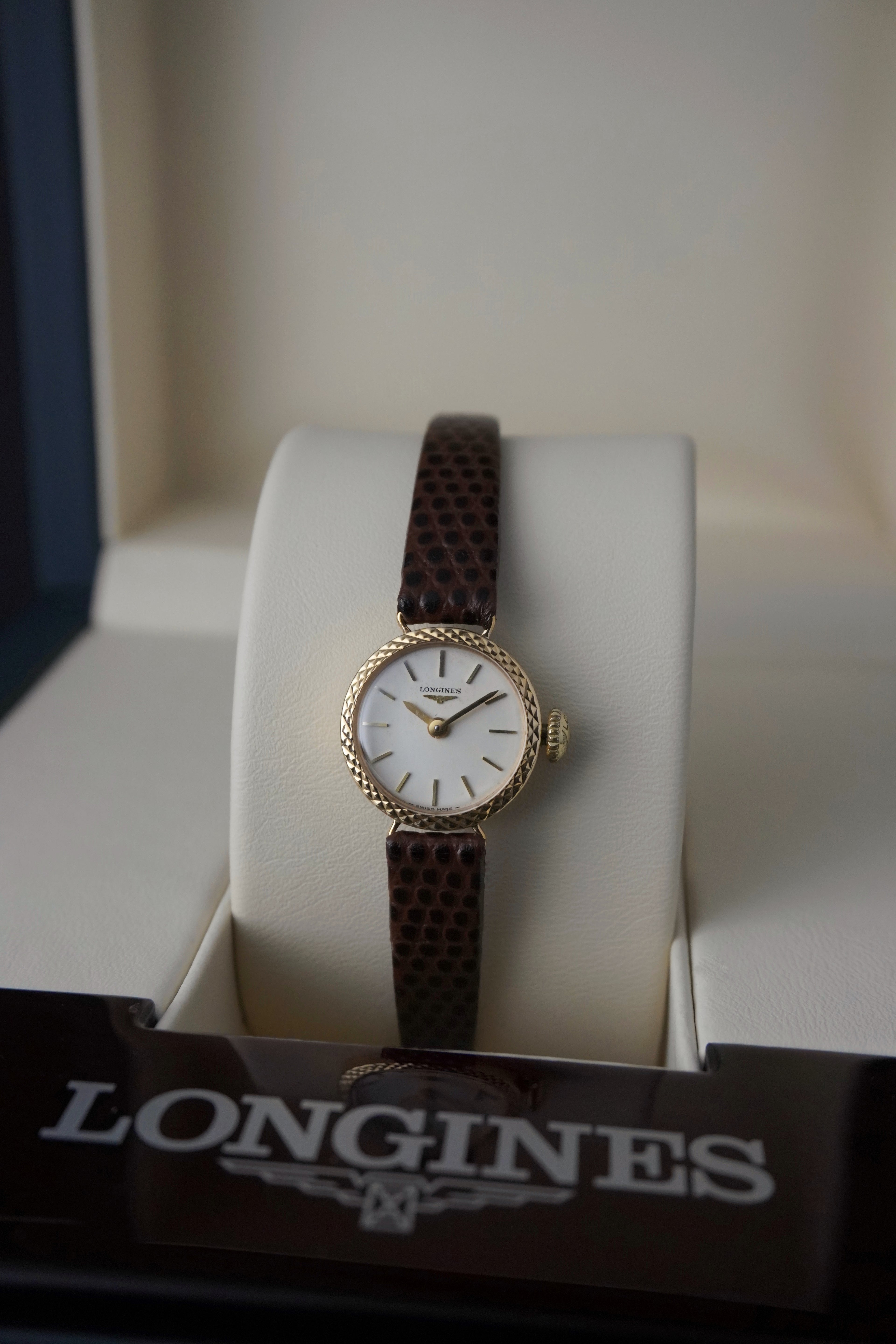 
Vintage Longines ladies’ wristwatch with a textured gold round case, white dial, slim gold hour markers, and brown leather strap, displayed on a cream watch cushion inside a Longines presentation box.