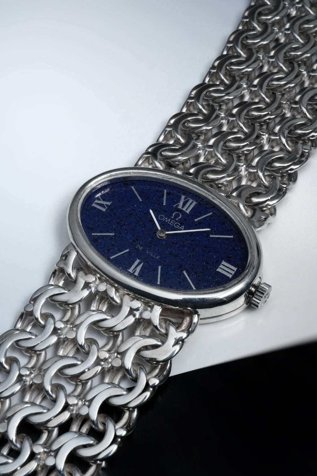 Solid silver ladies Omega De Ville watch with oval case, deep blue textured dial, Roman numerals, and integrated wide silver chain bracelet.