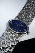 Solid silver ladies Omega De Ville watch with oval case, deep blue textured dial, Roman numerals, and integrated wide silver chain bracelet.