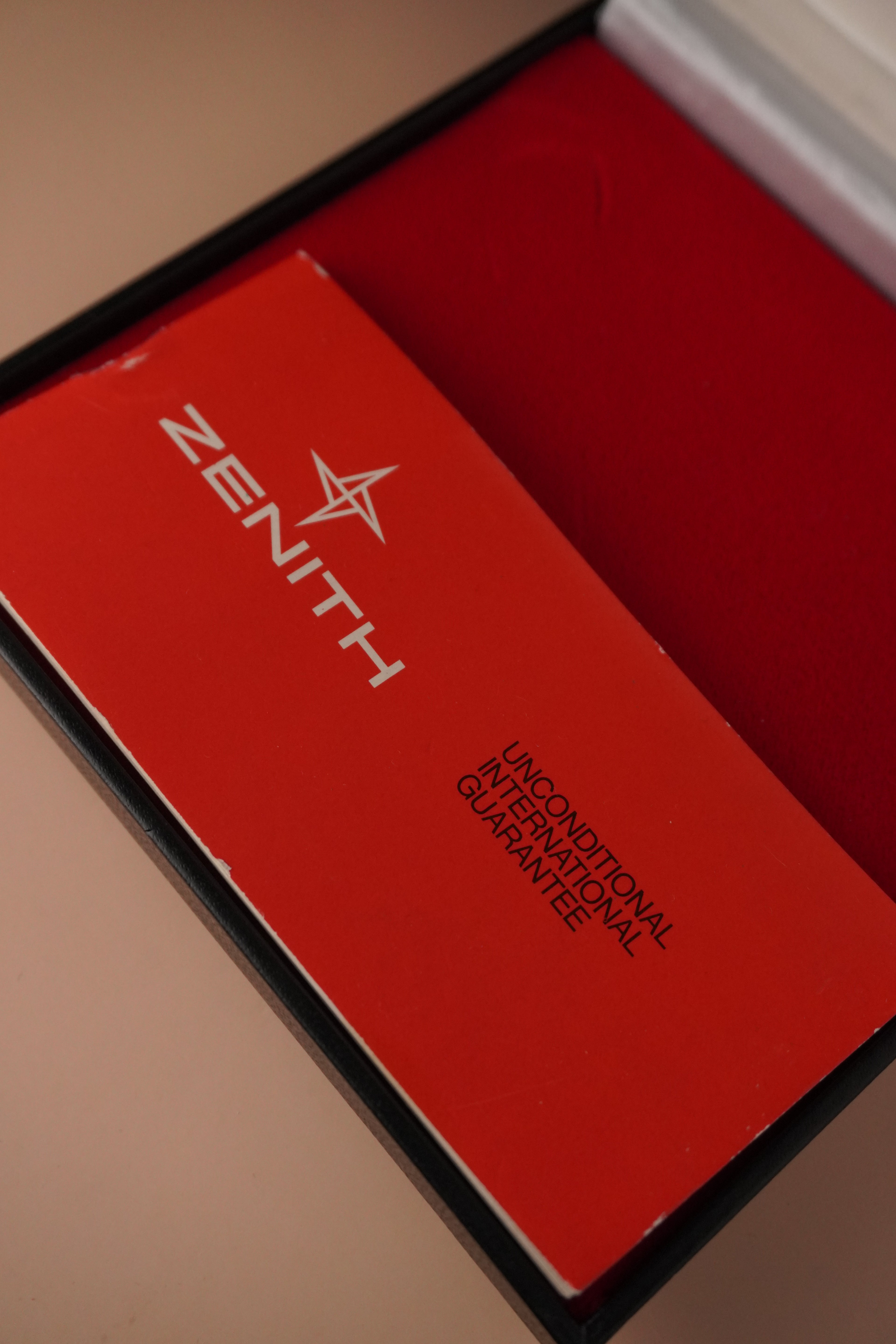 Zenith vintage dress watch with 9ct English-made yellow gold case, silver dial, black leather strap, professionally polished, displayed with original Zenith booklet on red background.