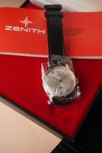 Zenith vintage dress watch with 9ct English-made yellow gold case, silver dial, black leather strap, professionally polished, displayed with original Zenith booklet on red background.