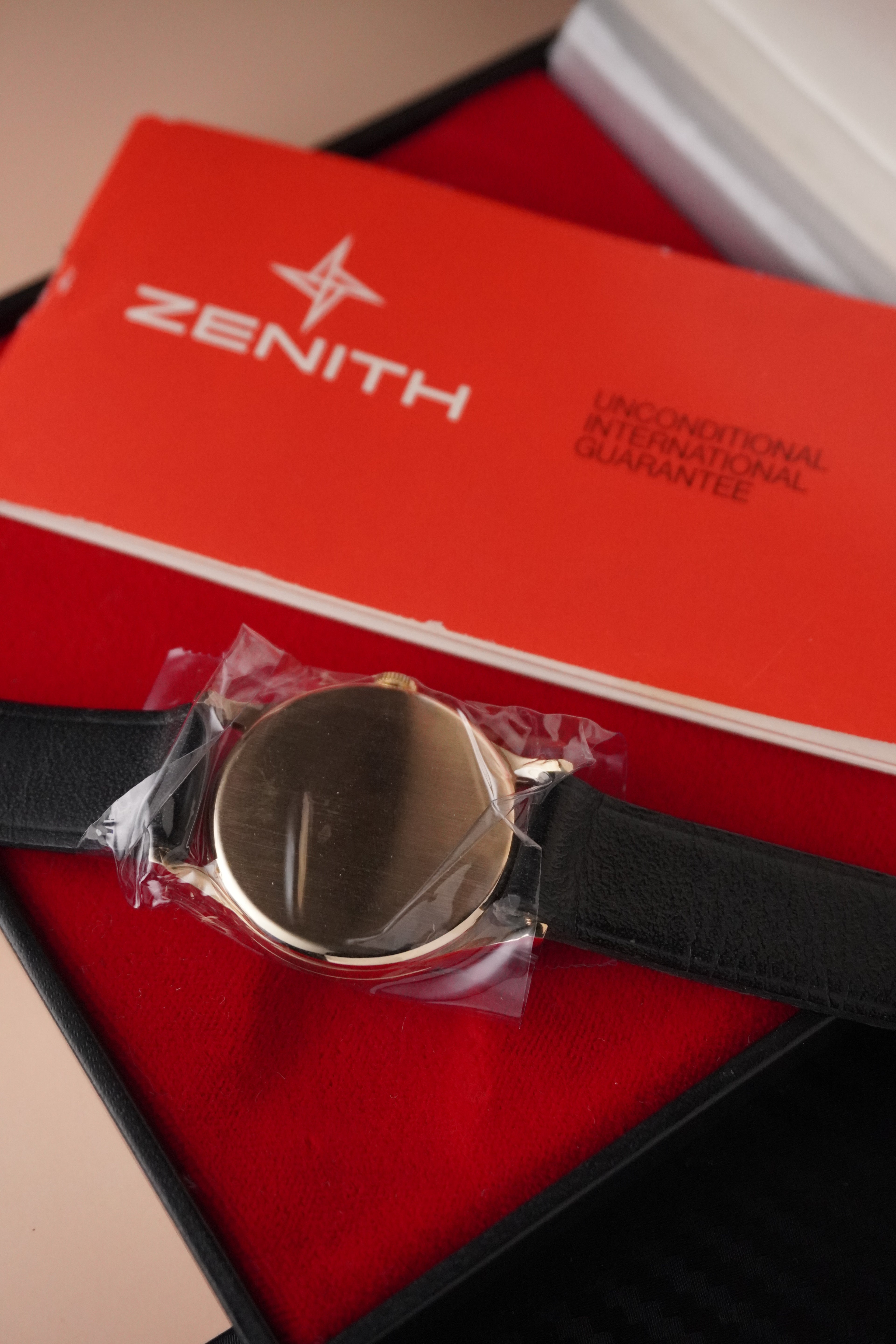 Zenith vintage dress watch with 9ct English-made yellow gold case, silver dial, black leather strap, professionally polished, displayed with original Zenith booklet on red background.