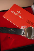 Gold watch with black strap and 'ZENITH' branding on a red cushion