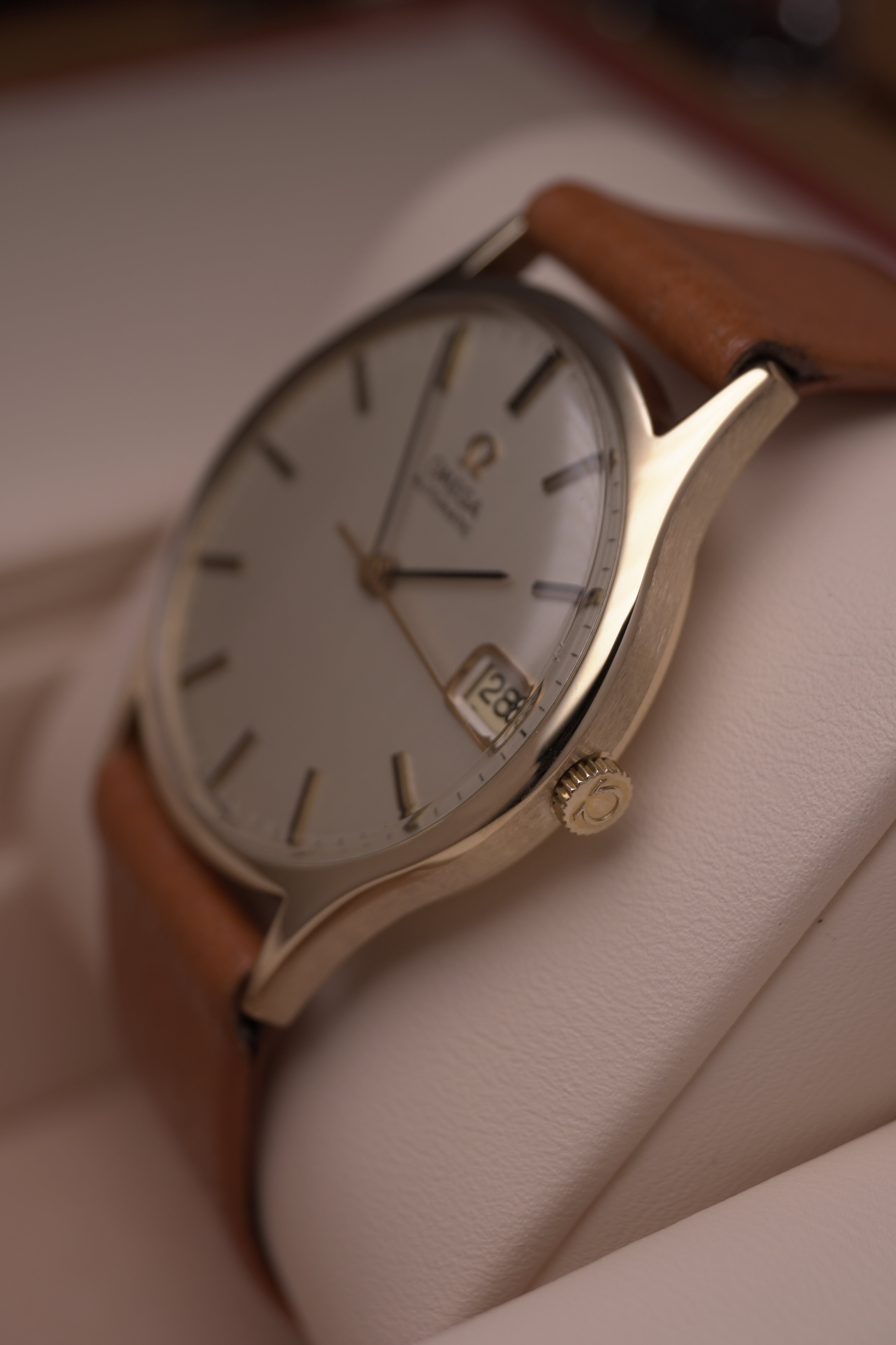 Angled close-up of a vintage Omega Automatic gents watch in solid yellow gold with a champagne dial, baton hour markers and date window, on a brown leather strap.”