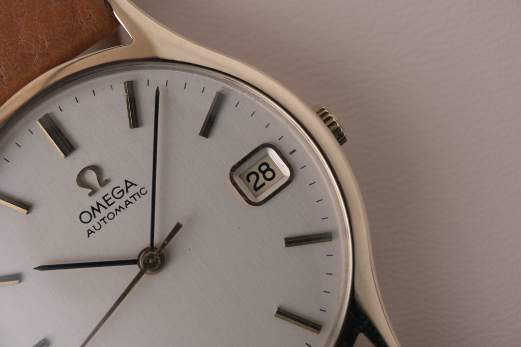 Angled close-up of a vintage Omega Automatic gents watch in solid yellow gold with a champagne dial, baton hour markers and date window, on a brown leather strap.