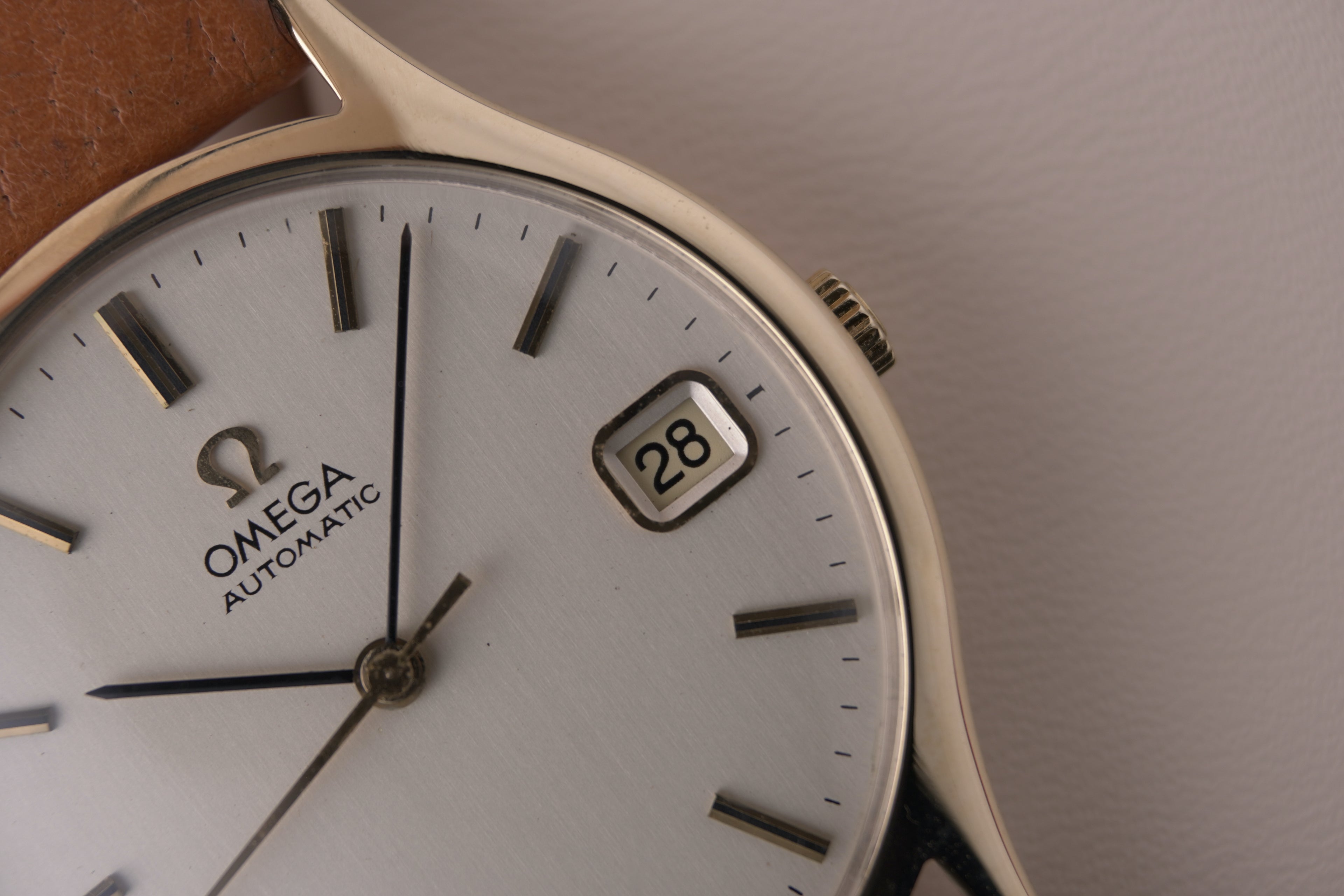 Angled close-up of a vintage Omega Automatic gents watch in solid yellow gold with a champagne dial, baton hour markers and date window, on a brown leather strap.