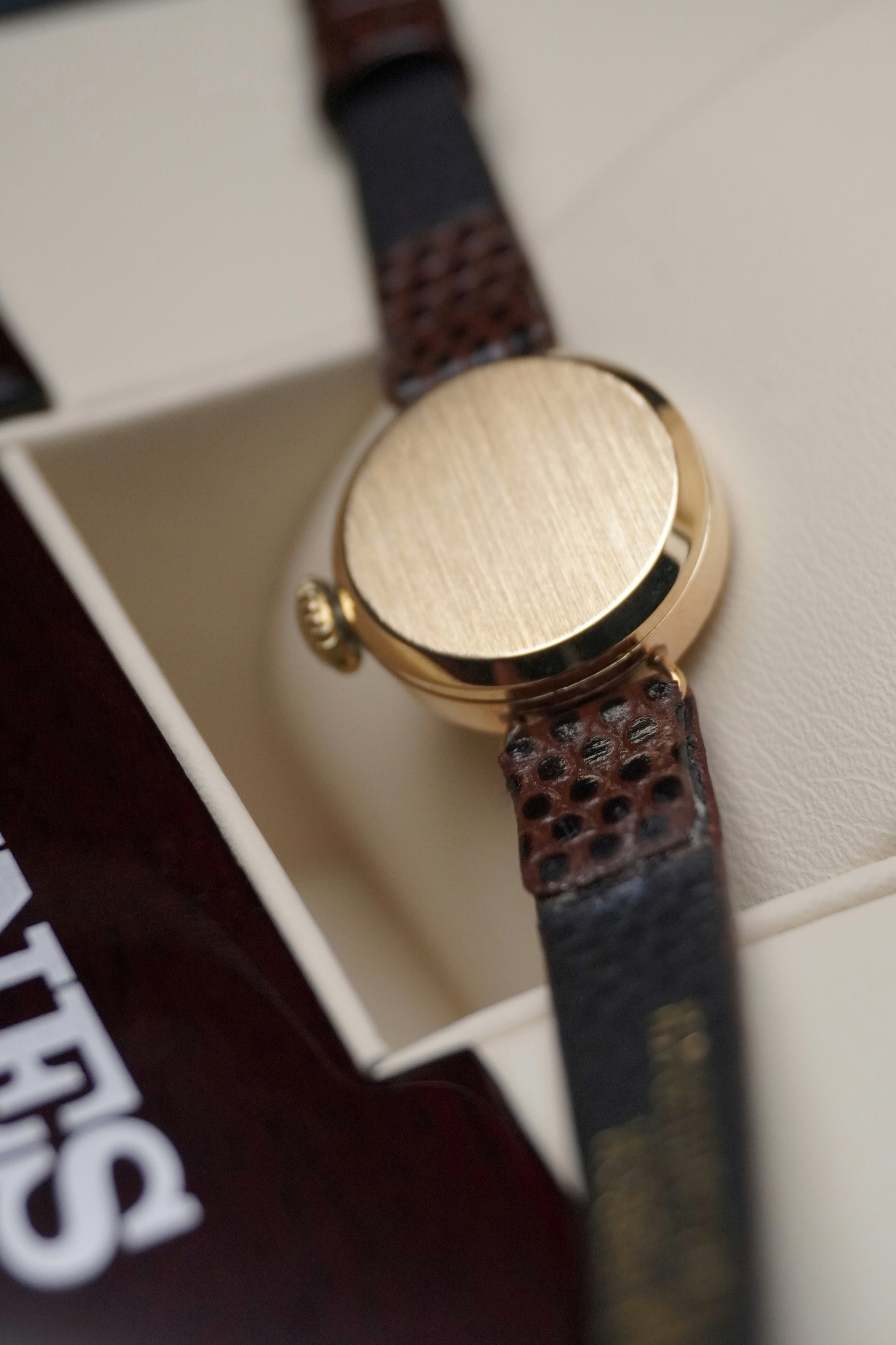 
Vintage Longines ladies’ wristwatch with a textured gold round case, white dial, slim gold hour markers, and brown leather strap, displayed on a cream watch cushion inside a Longines presentation box.